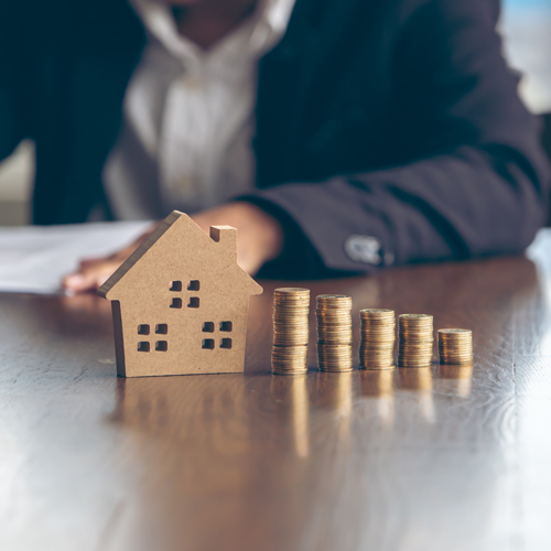Small wooden house on a desk with small stacks of coins cascading to the right.