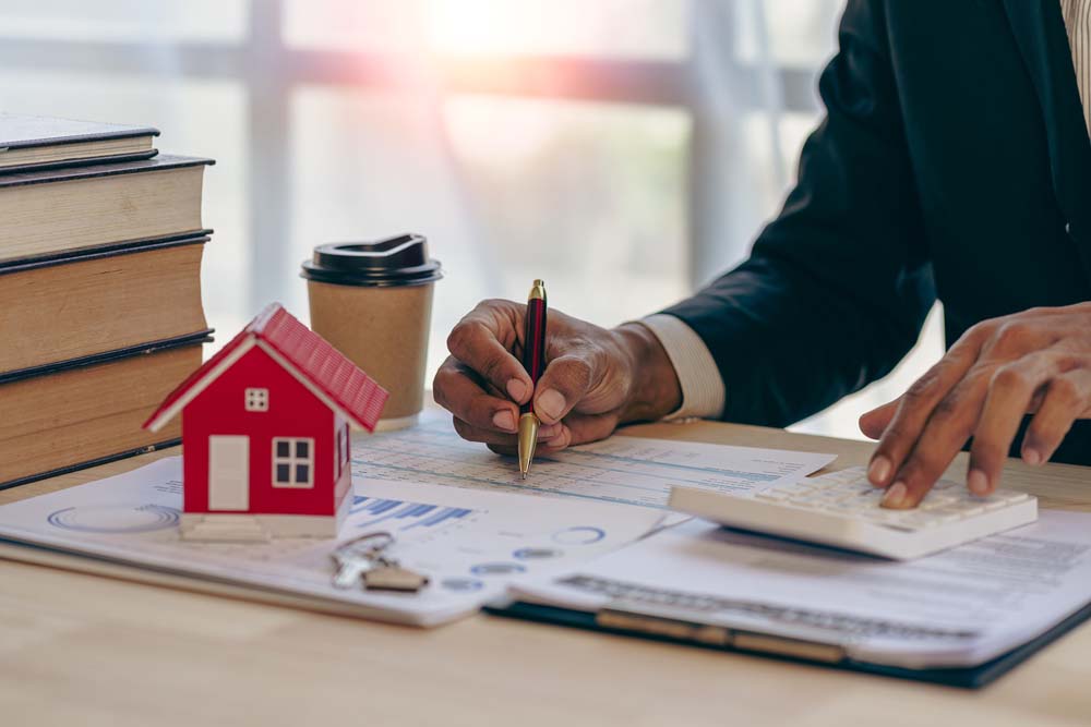 Man working at a desk with large books, coffee, spreadsheets, a clipboard, and a miniature, red model house with a set of keys beside it - equitable distribution and offsets