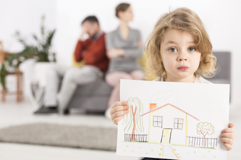 Upset little boy holding a drawing of a house with fighting parents in background.
