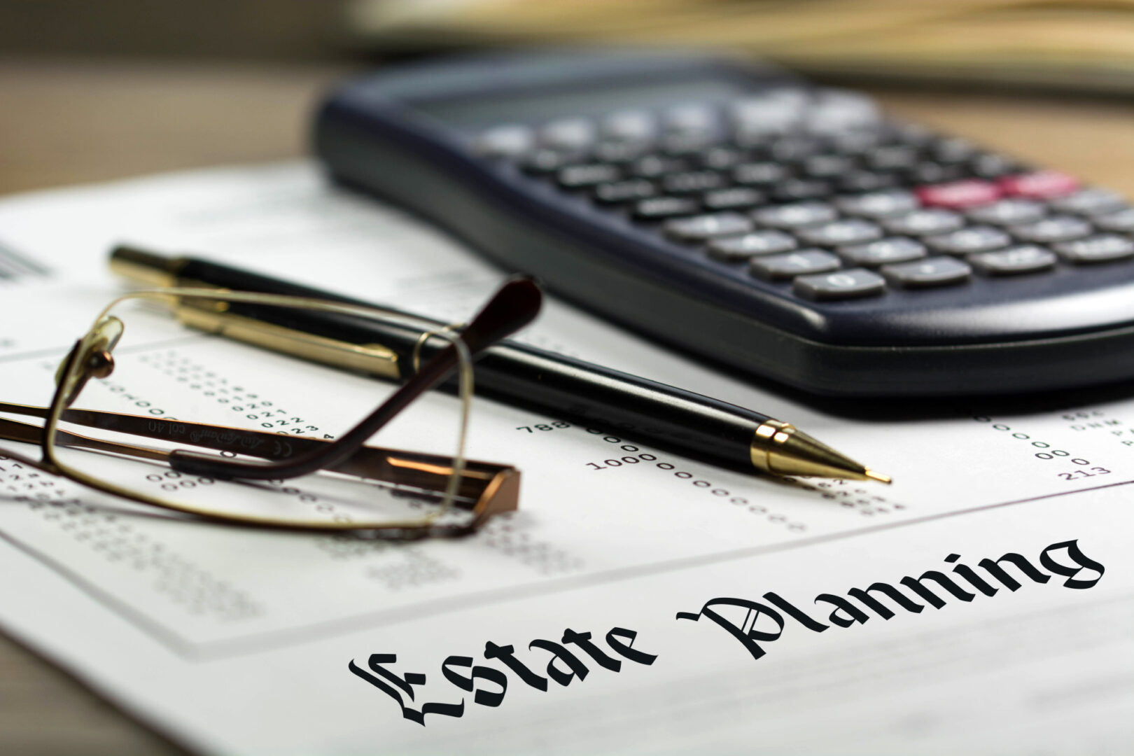 Close up of reading glasses, pen, and calculator sitting onto of paper that says Estate Planning on it