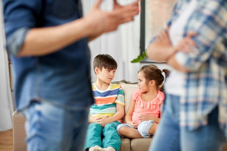 Cute girl and boy 4-5 years old sitting on couch and looking at each other with upset looks while faded image of parents are arguing