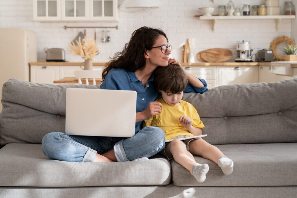 Loving mother businesswoman happy to work from home on coronavirus quarantine hug small son playing on digital tablet sit together with working remotely on laptop computer mom on sofa in living room