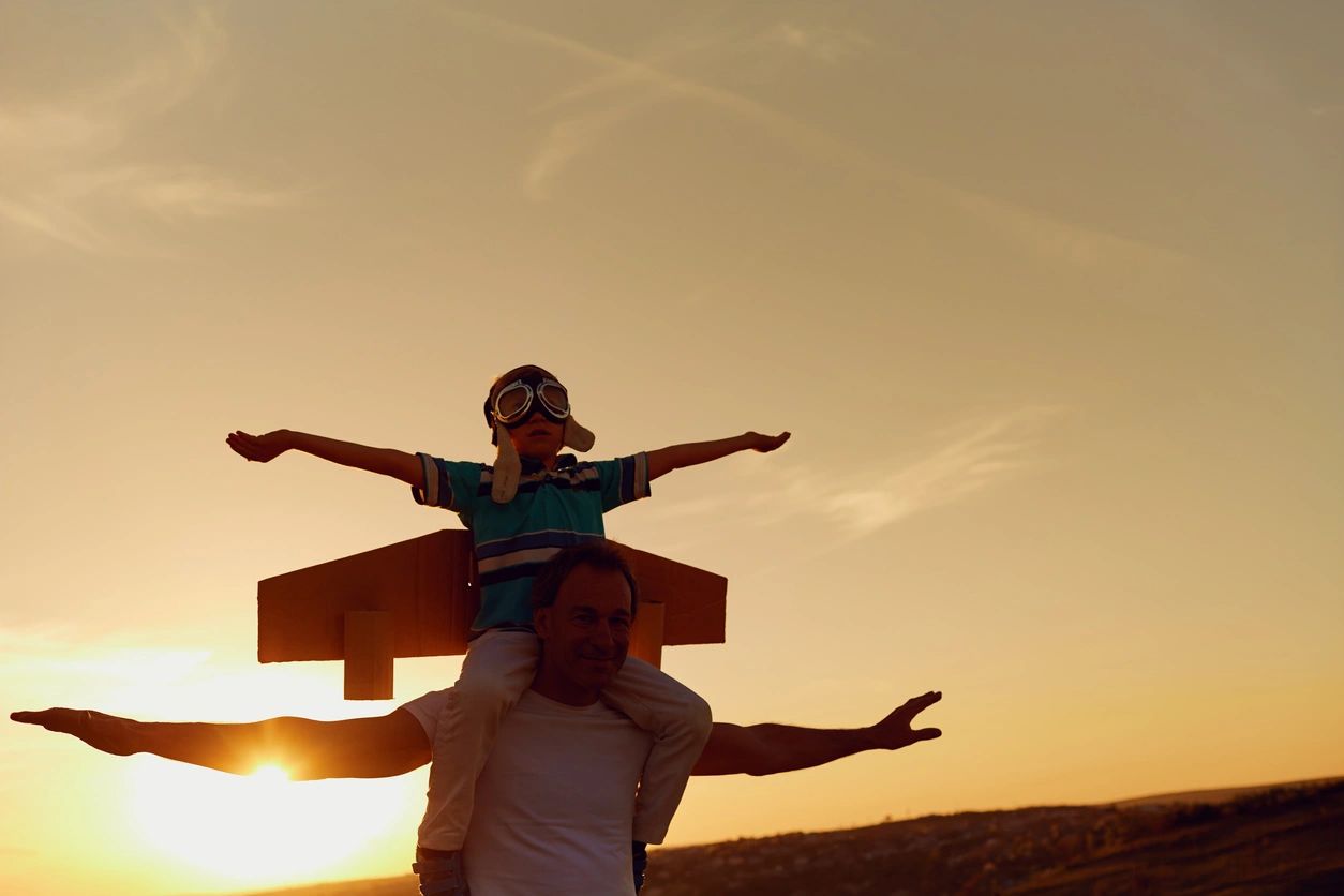 step father with step son on his shoulders, both with hands out to mimic an airplane